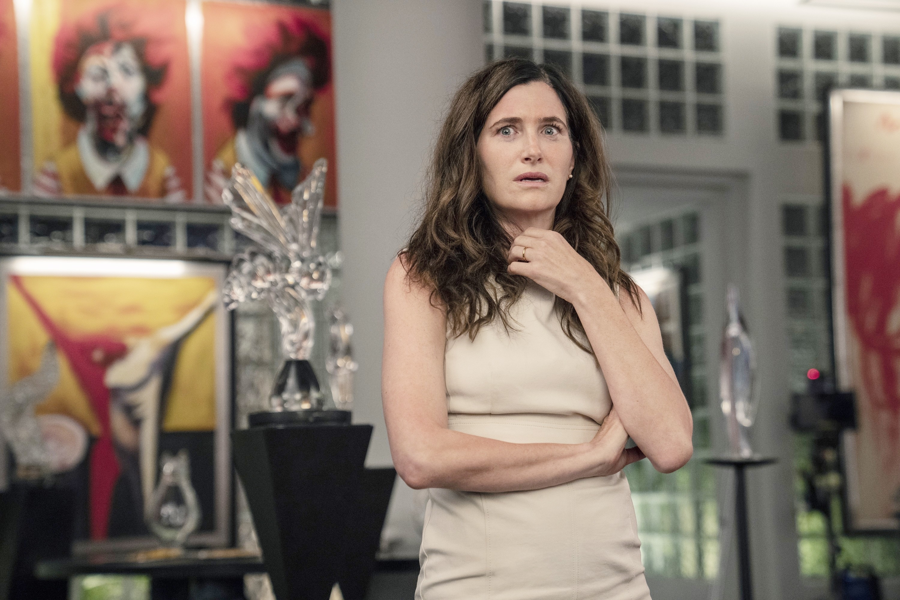 Kathryn Hahn stands in an art gallery wearing a sleeveless, high-neck dress, looking contemplative. Several abstract paintings and sculptures are in the background