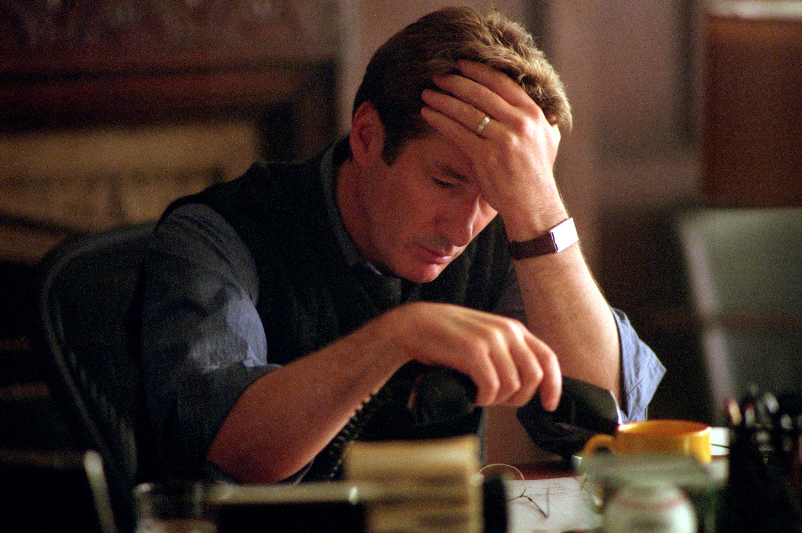 Richard Gere sits at a desk, looking stressed with his hand on his forehead, holding a phone. A yellow mug and documents are on the desk