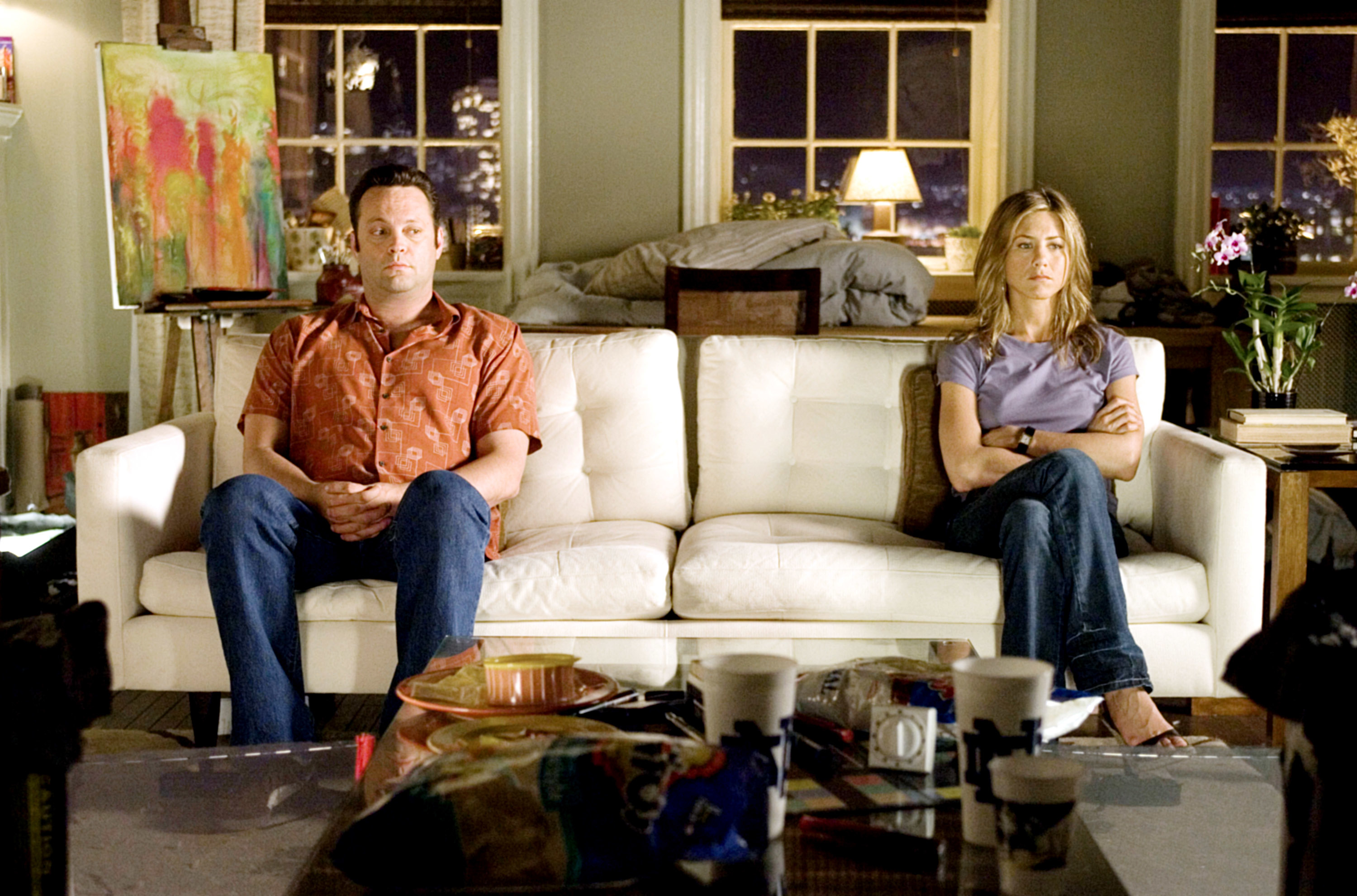 Vince Vaughn in a casual shirt and jeans, and Jennifer Aniston in a casual top and jeans, are sitting on a couch in a living room, appearing distant from each other
