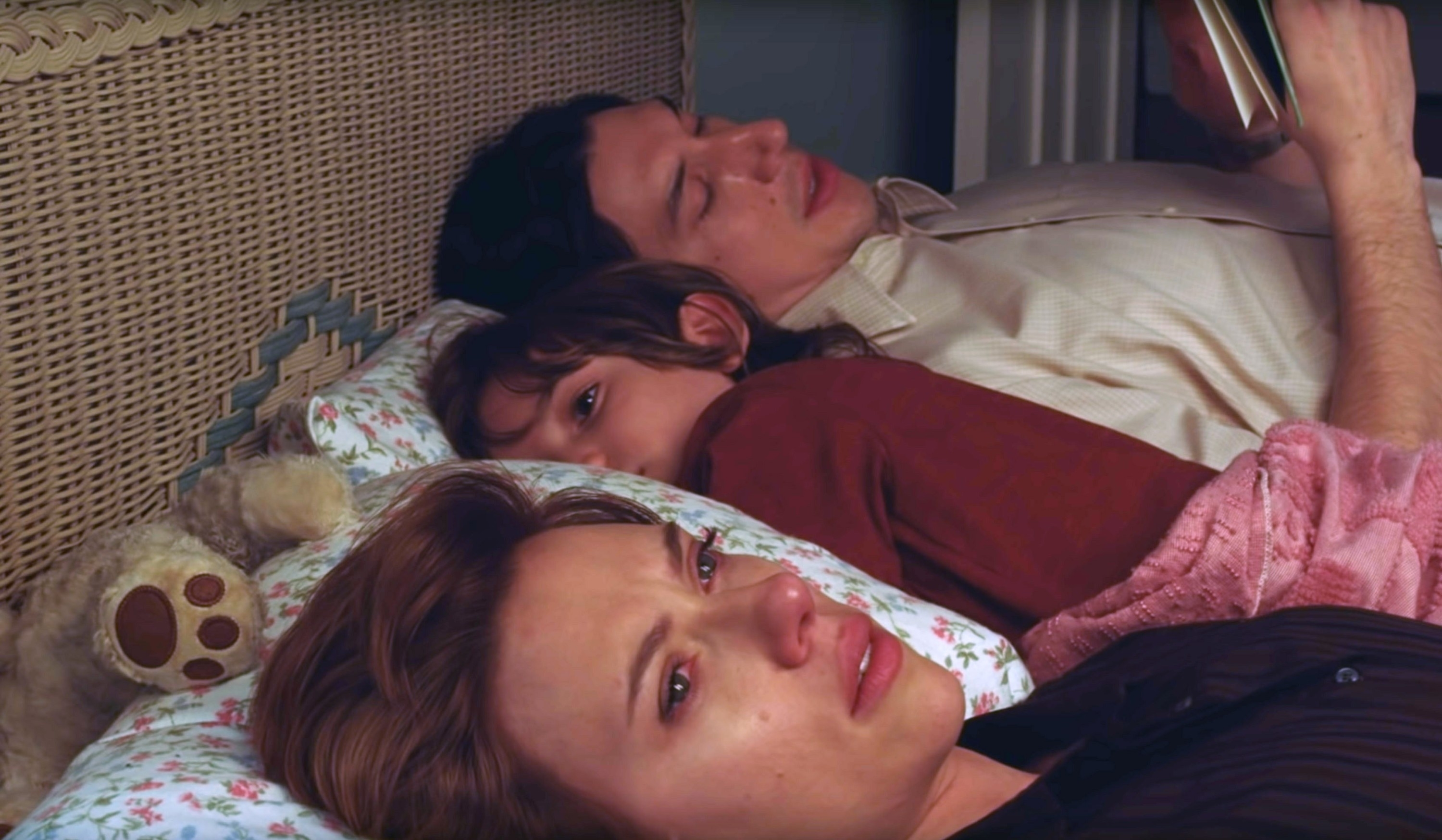 Scarlett Johansson, Adam Driver, and a child lie in bed. Scarlett is in the foreground, looking emotional. A teddy bear lies near the child and Adam is reading a book