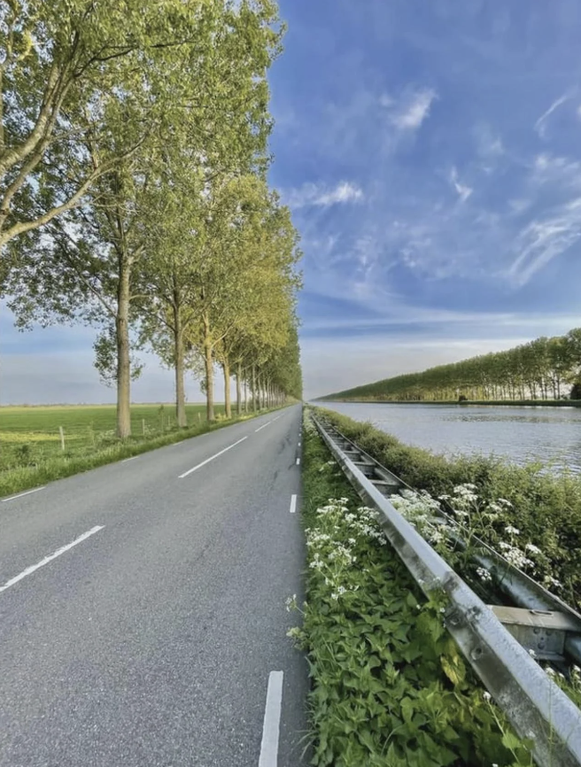 A long, empty road flanked by tall trees on the left and a calm river on the right, extending into the horizon under a partly cloudy sky