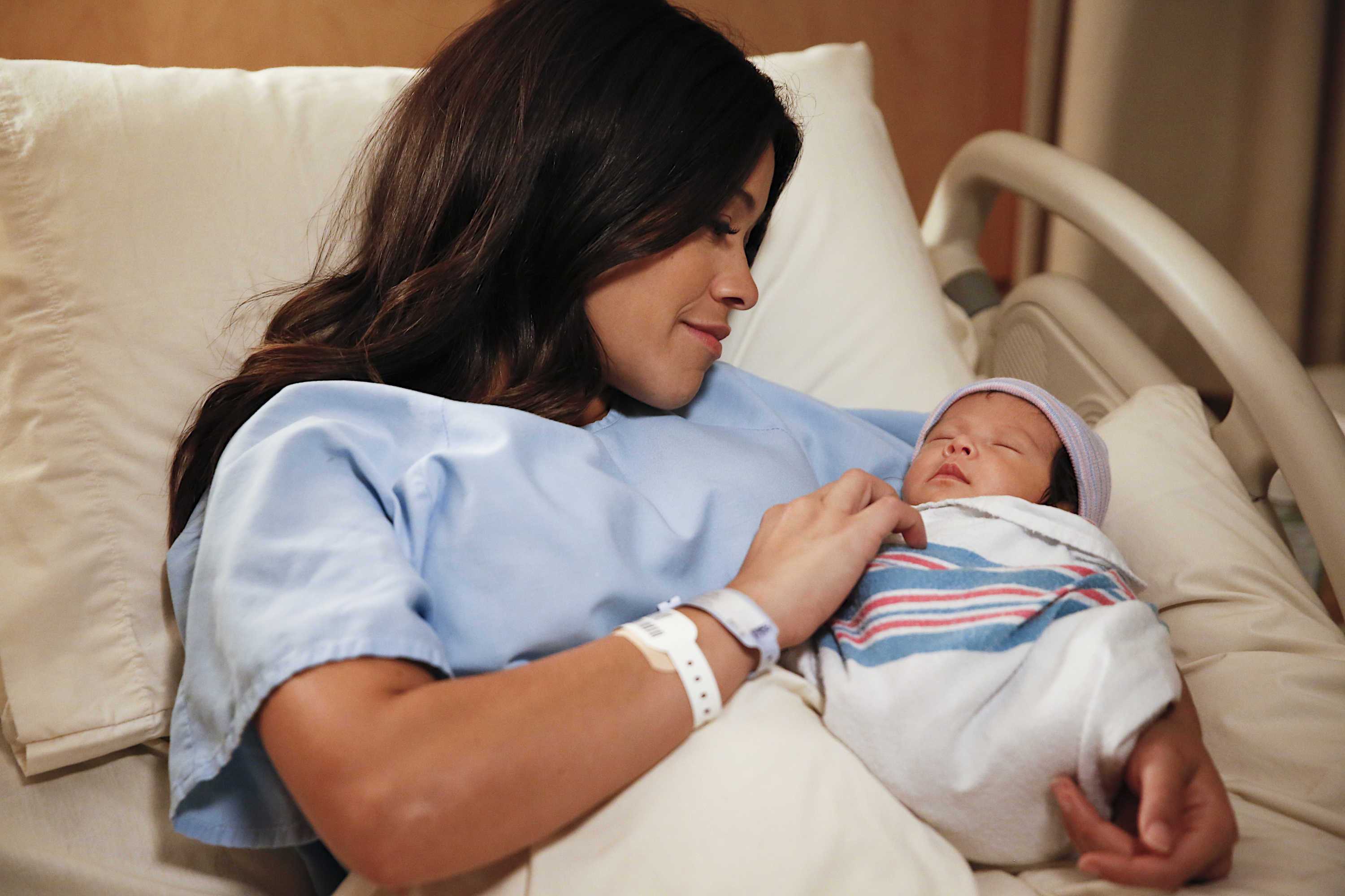 Gina Rodriguez is lying in a hospital bed, wearing a hospital gown, and smiling warmly while holding a newborn baby wrapped in a striped blanket