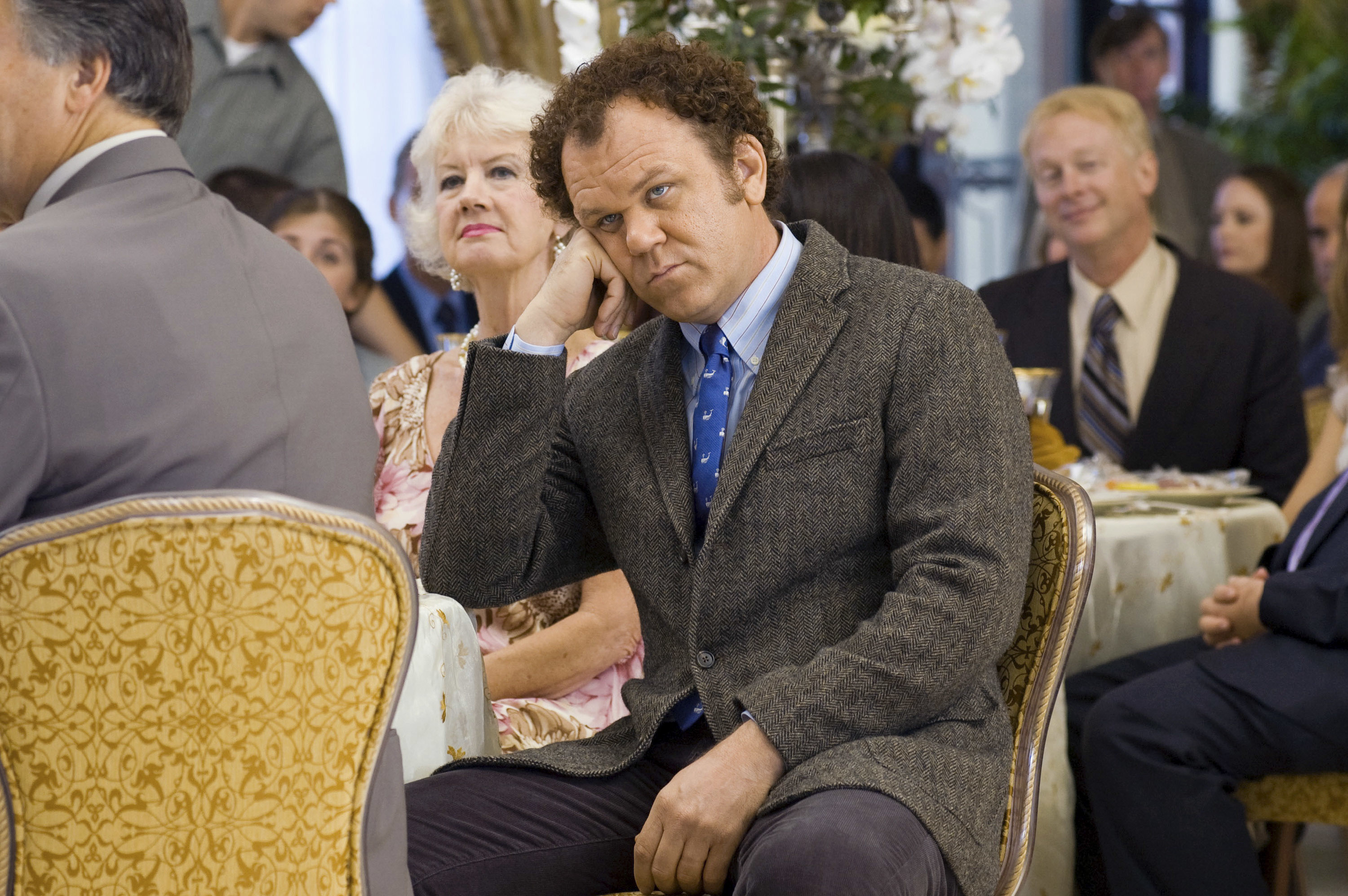 John C. Reilly, seated in a formal setting, looking directly at the camera while resting his head on his hand. Others in formal attire are in the background