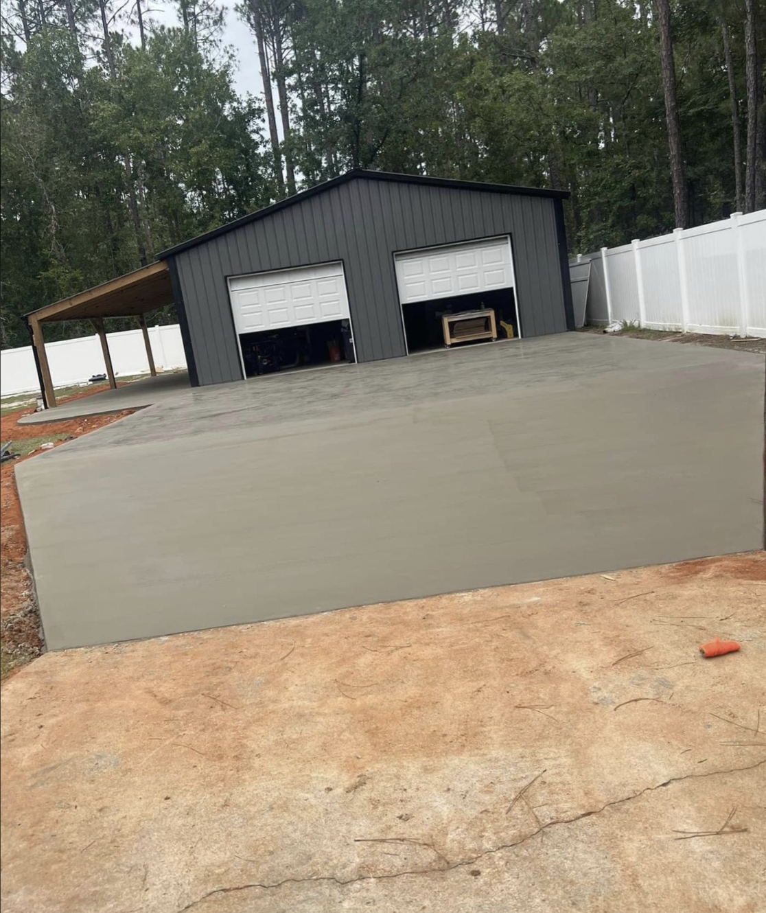 A newly constructed concrete driveway leads to a double-door garage with an adjacent carport in a wooded backyard setting