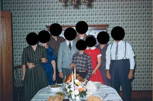 Eight people stand behind a table with a large meal, including a turkey, candles, and sides. They are in a dining room with patterned wallpaper