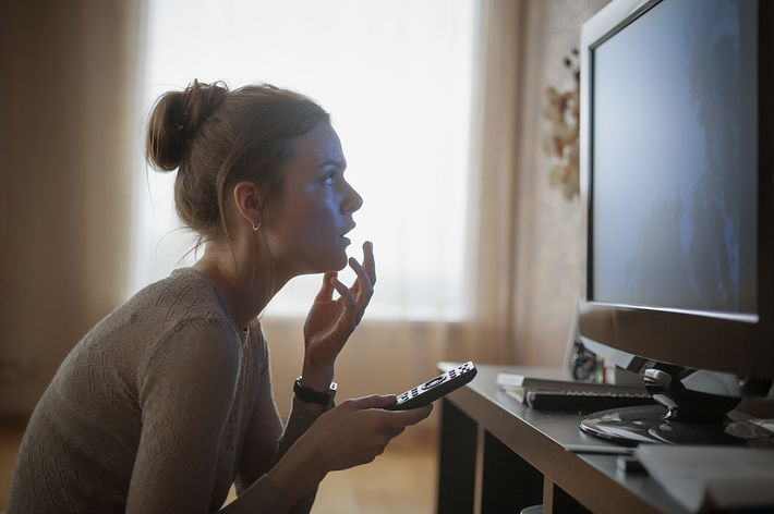 女性がテレビを見ている様子。リモコンを手に持ち、考え込むような表情を浮かべている。