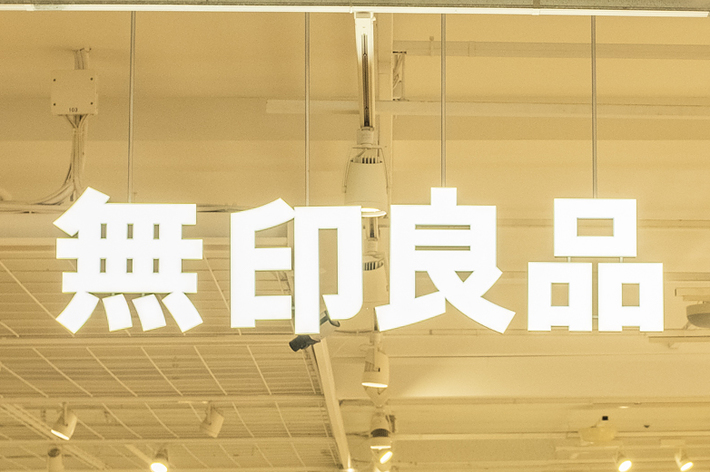 A store interior with shoppers walking around, featuring shelves filled with various items. The store sign has Japanese characters that translate to &ldquo;MUJI.&quot;