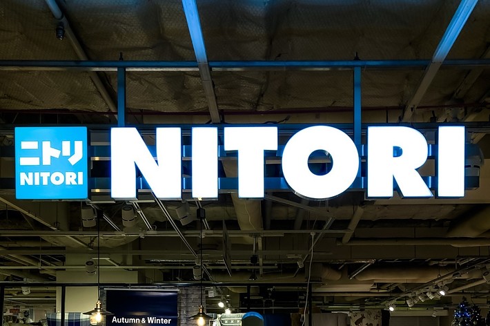 ニトリの店舗看板。ライトアップされた「NITORI」のロゴが表示されている。背景には秋冬コレクションの広告が見える。