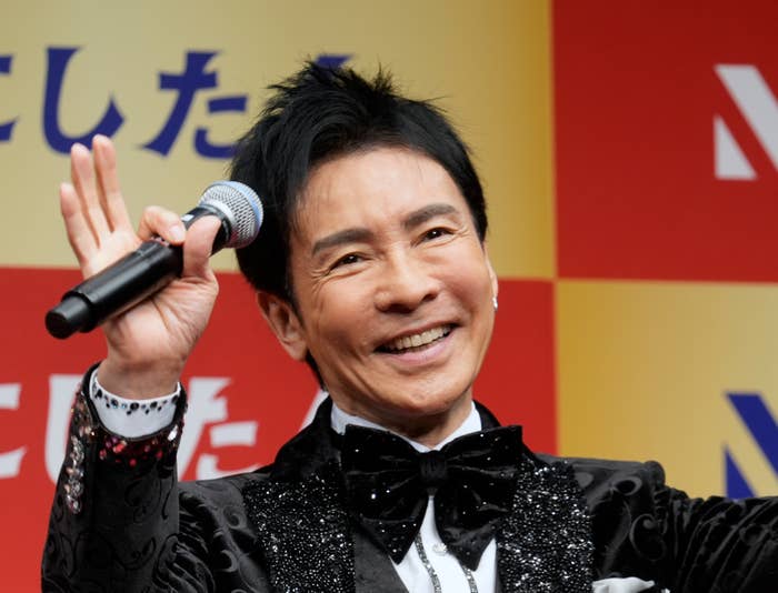 男性タレントがマイクを持って笑顔でポーズしている、豪華な服装。