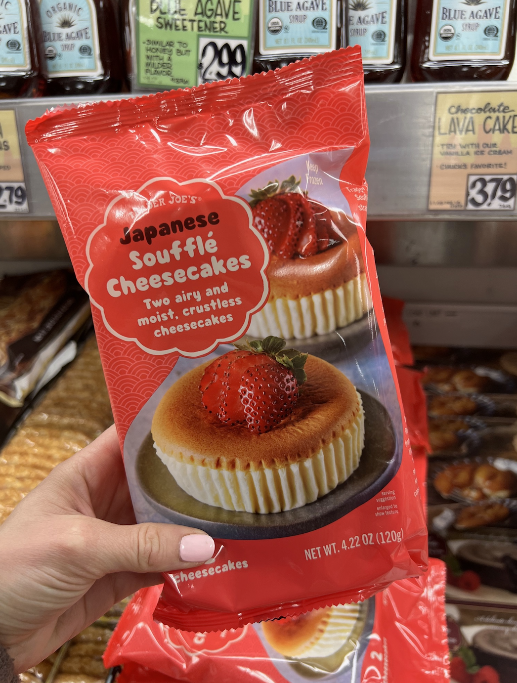 A person holds a package of Trader Joe's Japanese Soufflé Cheesecakes, described as "two airy and moist, crustless cheesecakes" in a grocery store aisle