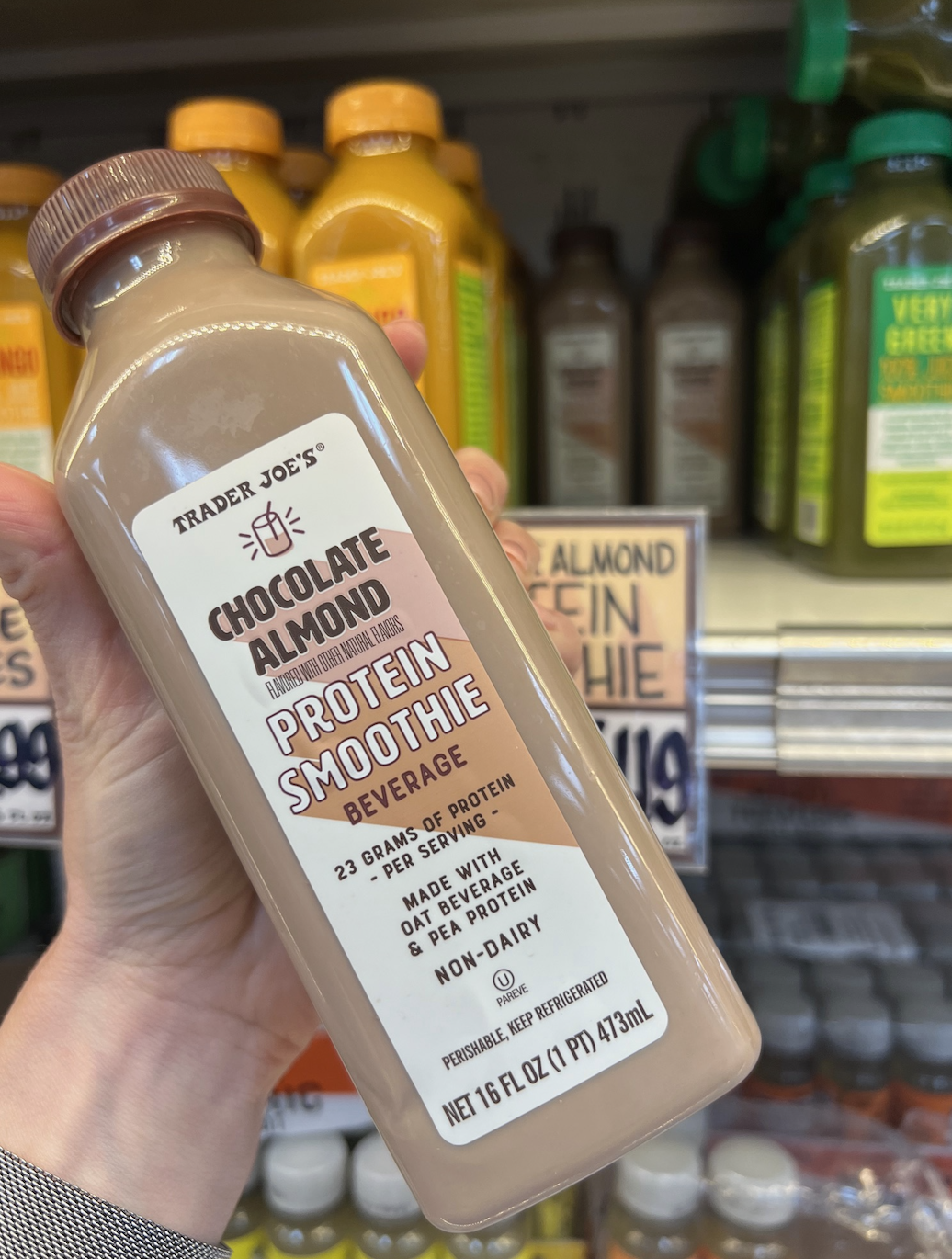 Hand holding a bottle of Trader Joe's Chocolate Almond Protein Smoothie beverage in front of store shelves stocked with similar drinks