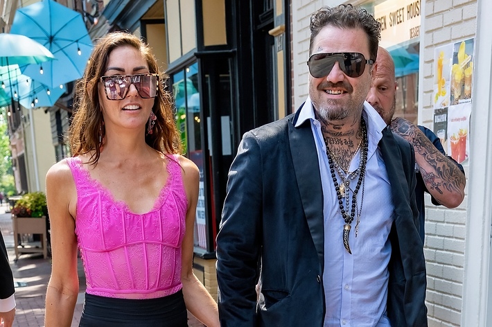 A man with tattoos and wearing sunglasses, a dark coat, and a white shirt walks beside a woman in a sleeveless top and sunglasses. Both are smiling