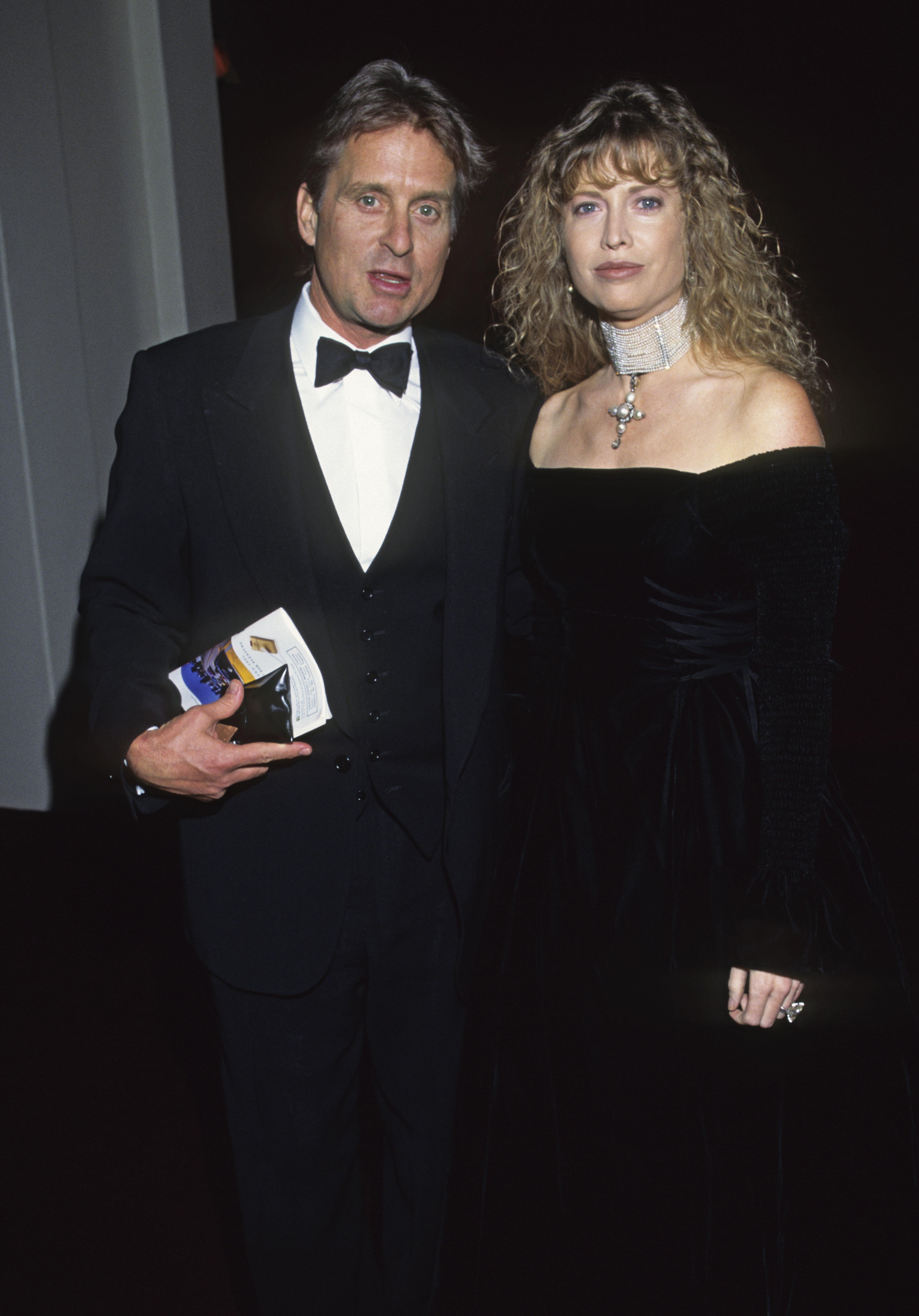 Michael Douglas in a tuxedo and Catherine Zeta-Jones in an off-the-shoulder dress with a choker necklace, posing for a photo