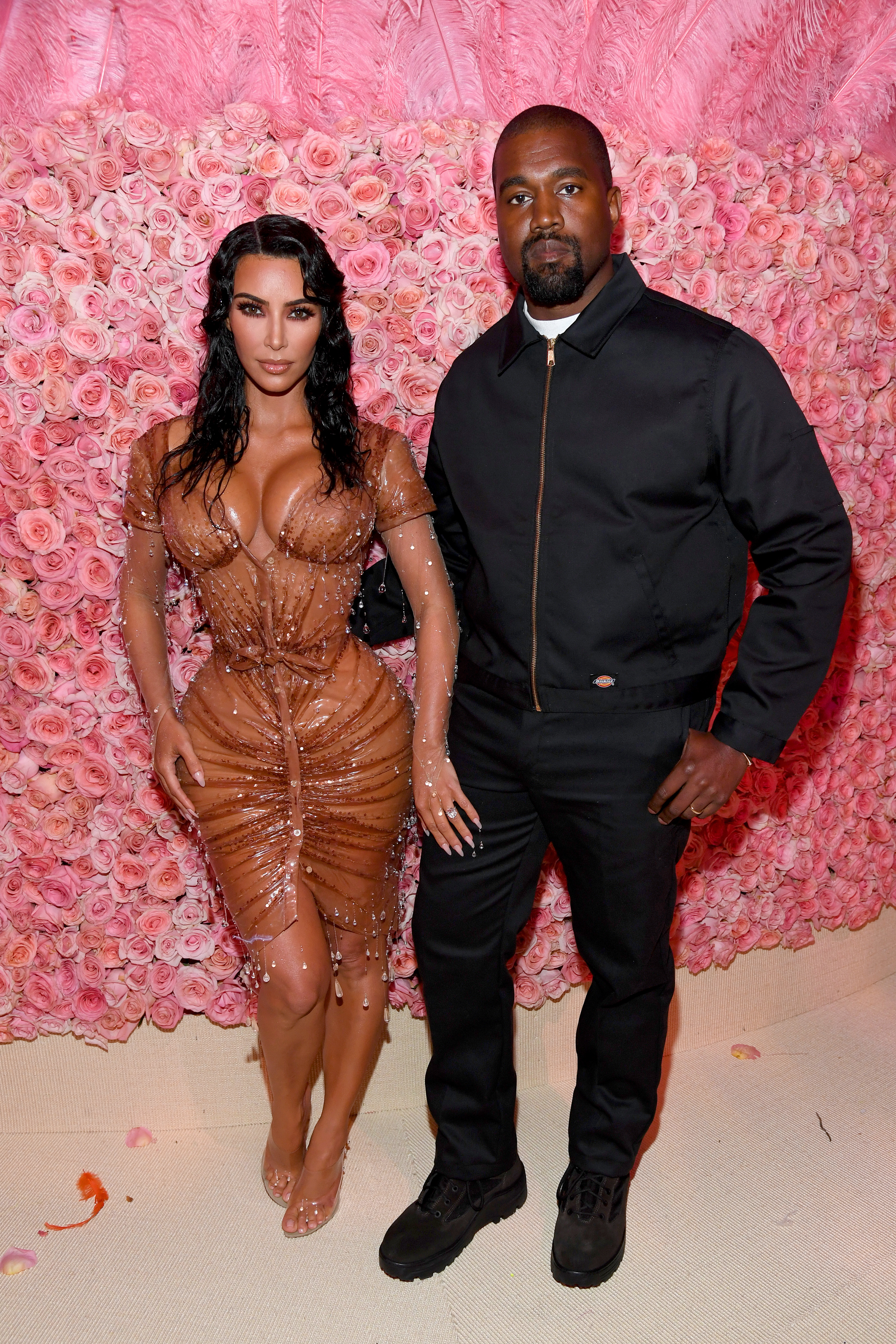 Kim Kardashian and Kanye West pose together, with Kim in a form-fitting, sheer dress and Kanye in a black jacket and pants, against a background of flowers