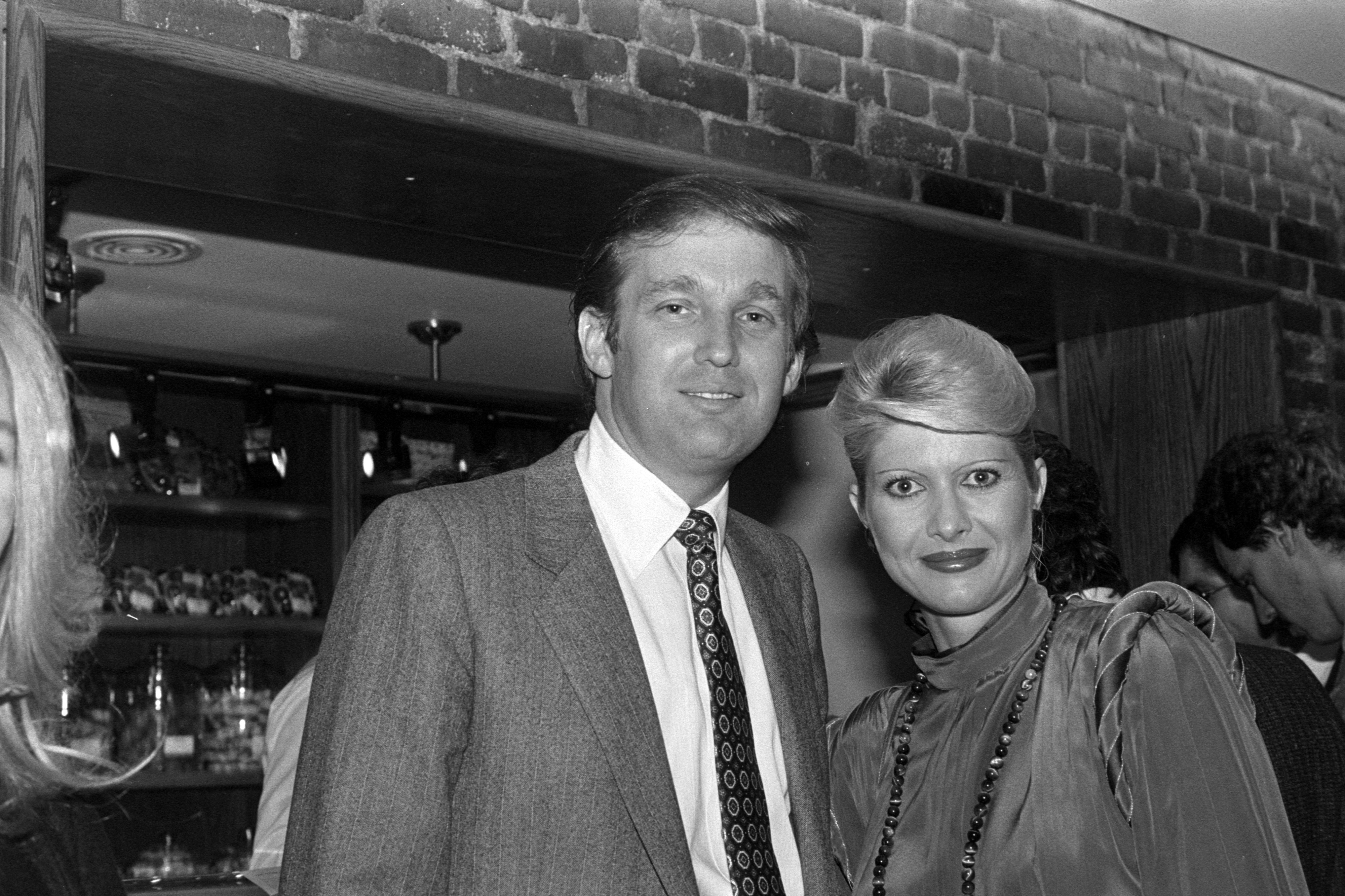 Donald Trump and Ivana Trump stand together, both smiling. Donald is in a suit and tie, while Ivana wears a stylish dress with a high collar and long necklace