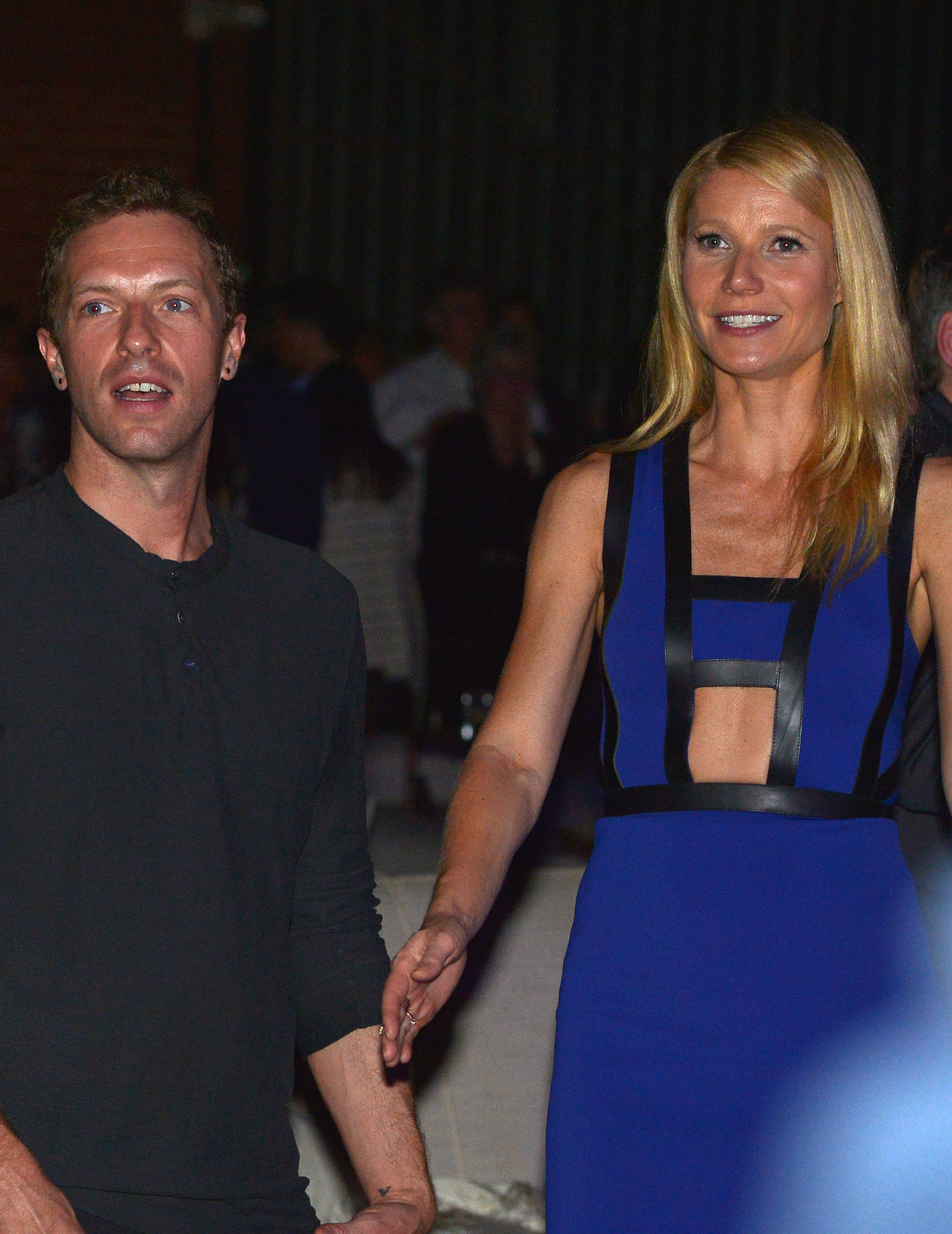 Chris Martin and Gwyneth Paltrow are holding hands; Chris is wearing a black shirt, and Gwyneth is in a sleeveless, stylish dress with cutout details