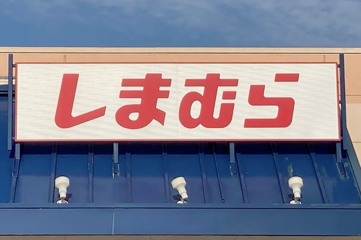 Storefront with Japanese signage reading &ldquo;しまむら&rdquo; (Shimamura) under a partly cloudy sky. The store entrance has a blue awning and accessible parking sign