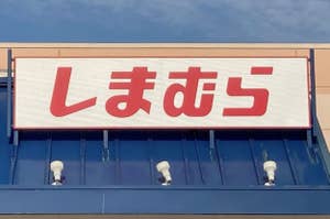 Storefront with Japanese signage reading "しまむら" (Shimamura) under a partly cloudy sky. The store entrance has a blue awning and accessible parking sign