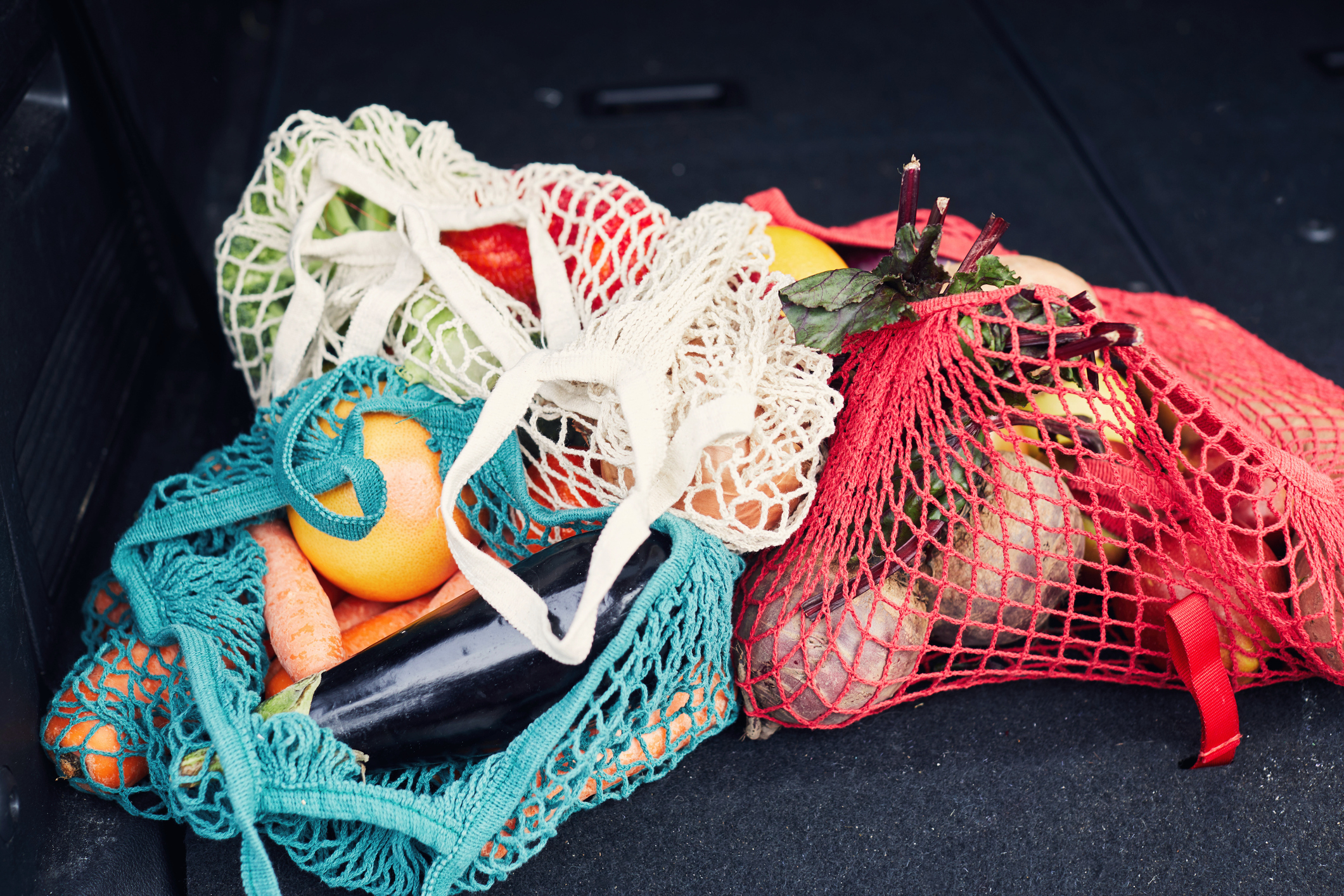 Three mesh bags filled with vegetables and fruits, including carrots, eggplants, orange, and greens, placed on a car trunk floor