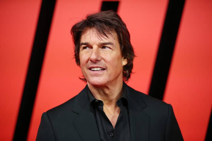 Tom Cruise on a red carpet, wearing a black suit and black shirt, smiling and looking slightly to the right