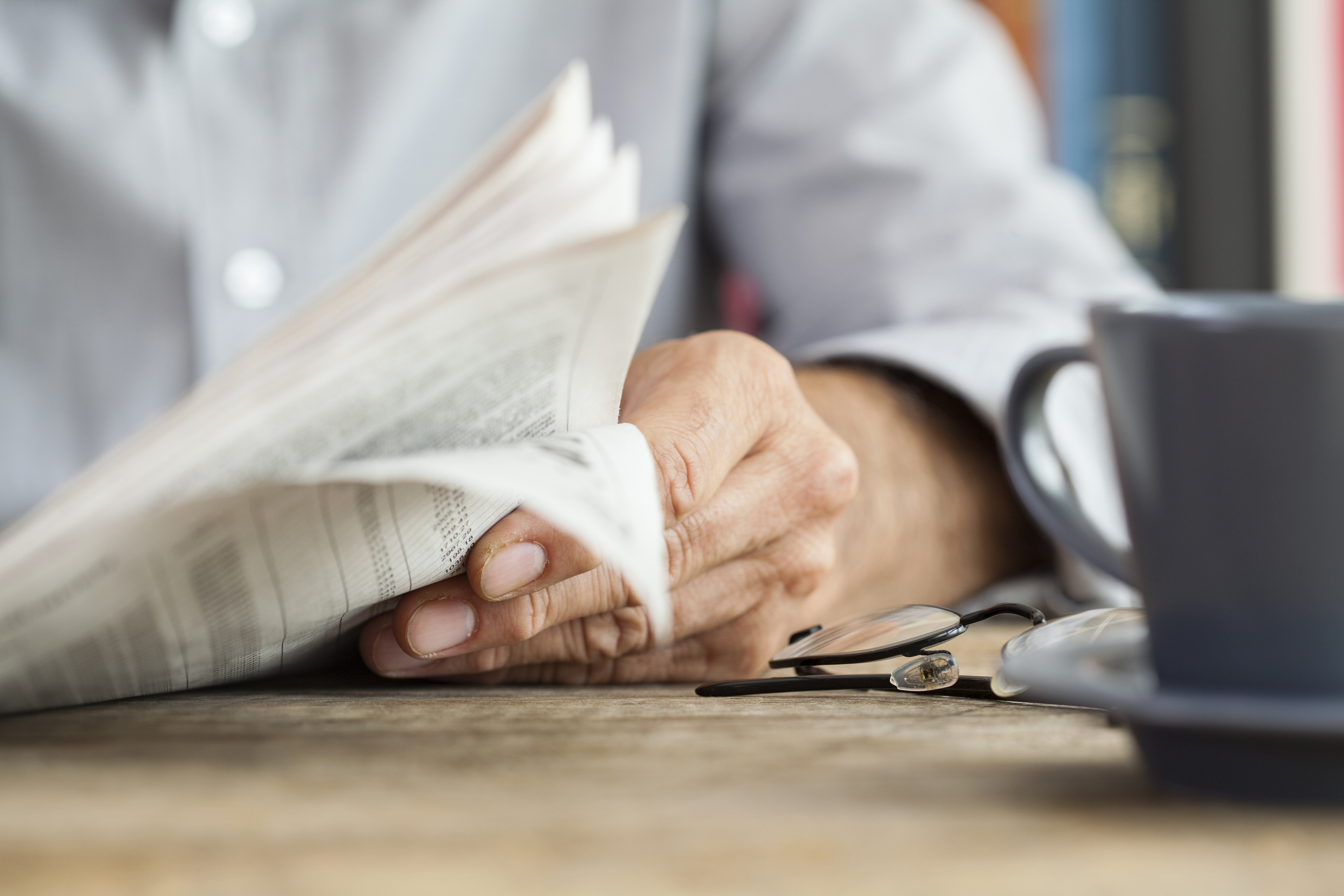 Close-up of a person reading a newspaper with a pair of glasses resting on a wooden table