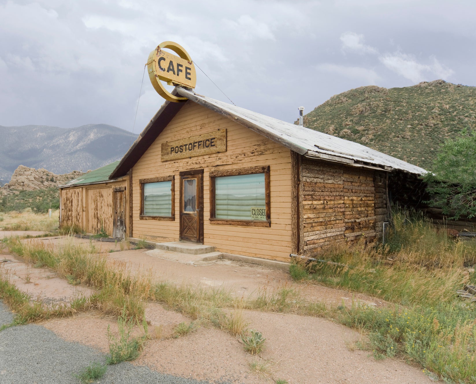 A rustic, wooden building with a "Cafe" sign and labeled "Post Office" appears closed. It is set against a mountain backdrop under a cloudy sky