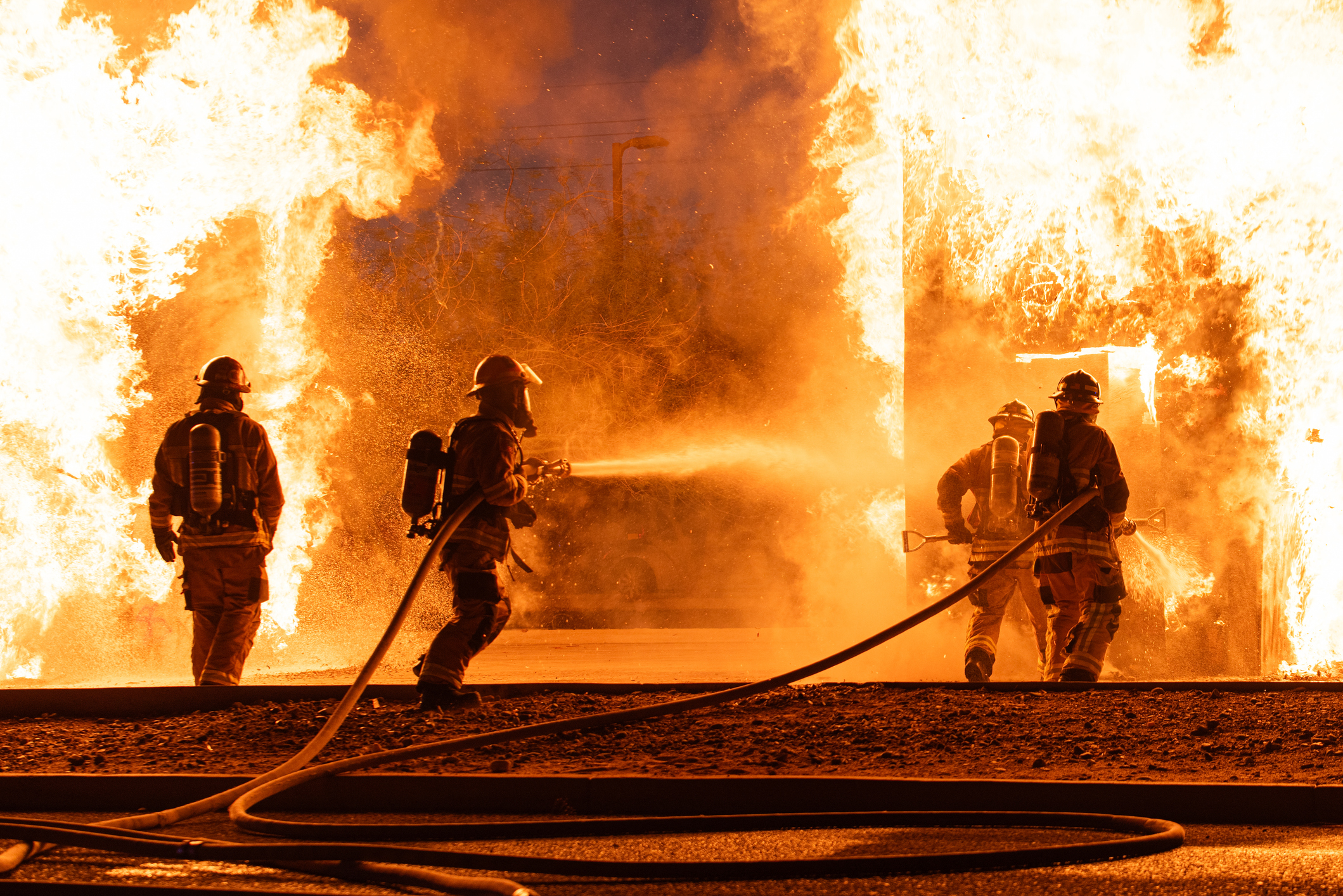 Three firefighters in full gear are battling a blazing fire with hoses, surrounded by intense flames