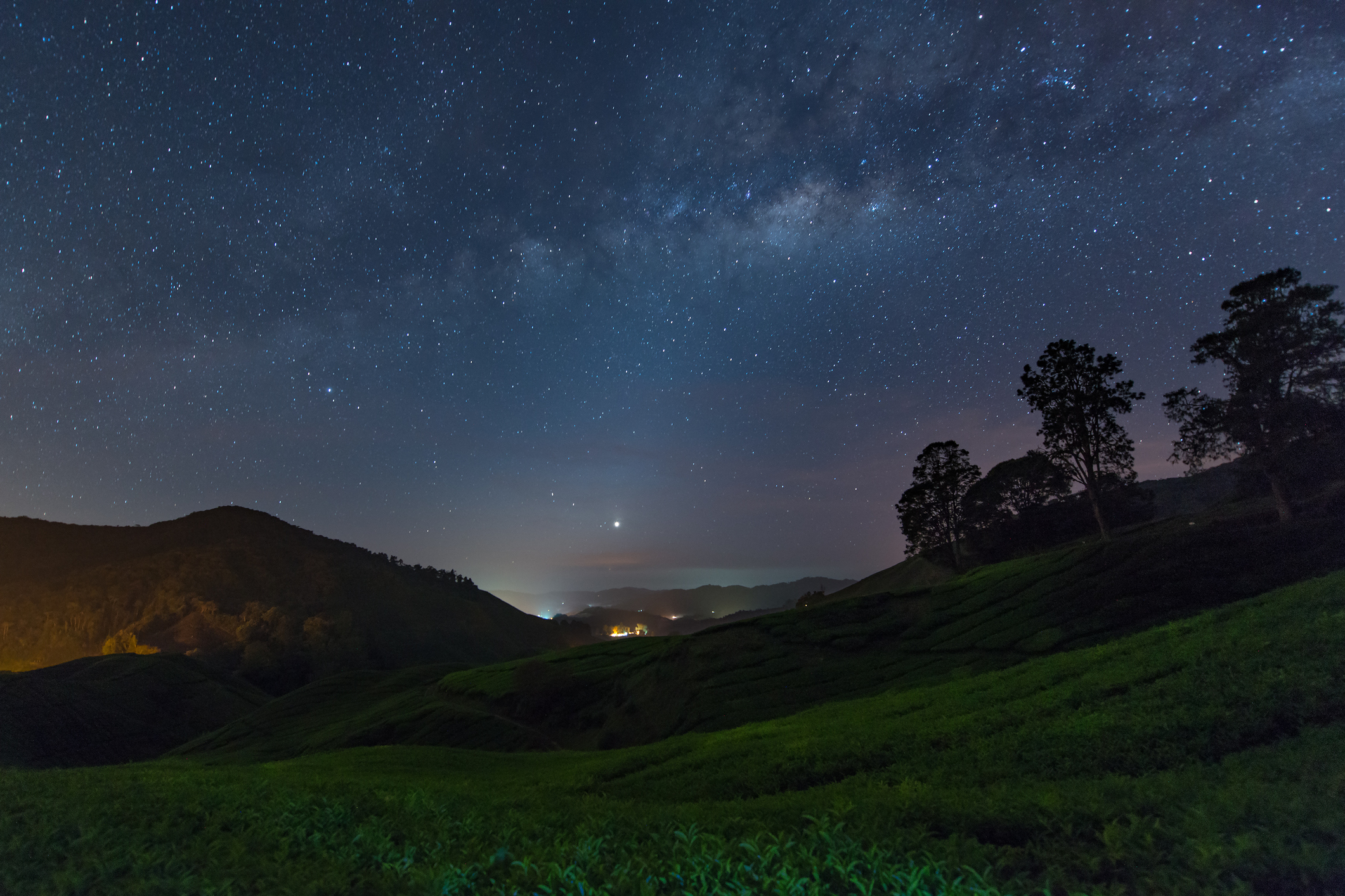 Nighttime landscape with a starry sky over rolling hills and a distant village illuminated by lights below
