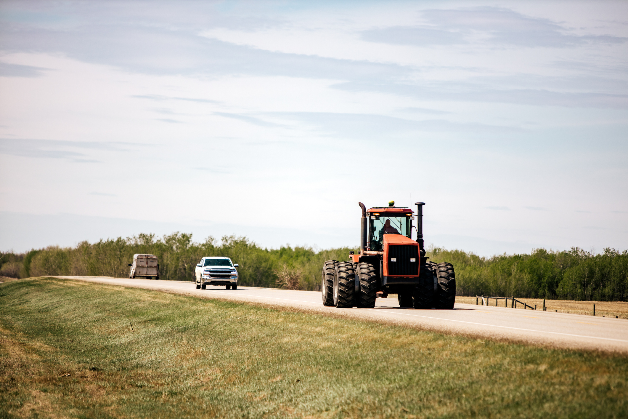 A tractor and two cars drive on a rural highway surrounded by fields and trees under a clear sky