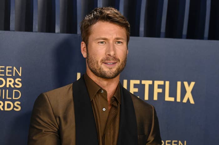 Glen Powell at an event, wearing a dark blazer over a button-up shirt, posing in front of a backdrop with text including &quot;Netflix&quot; and &quot;Screen Actors Guild Awards.&quot;