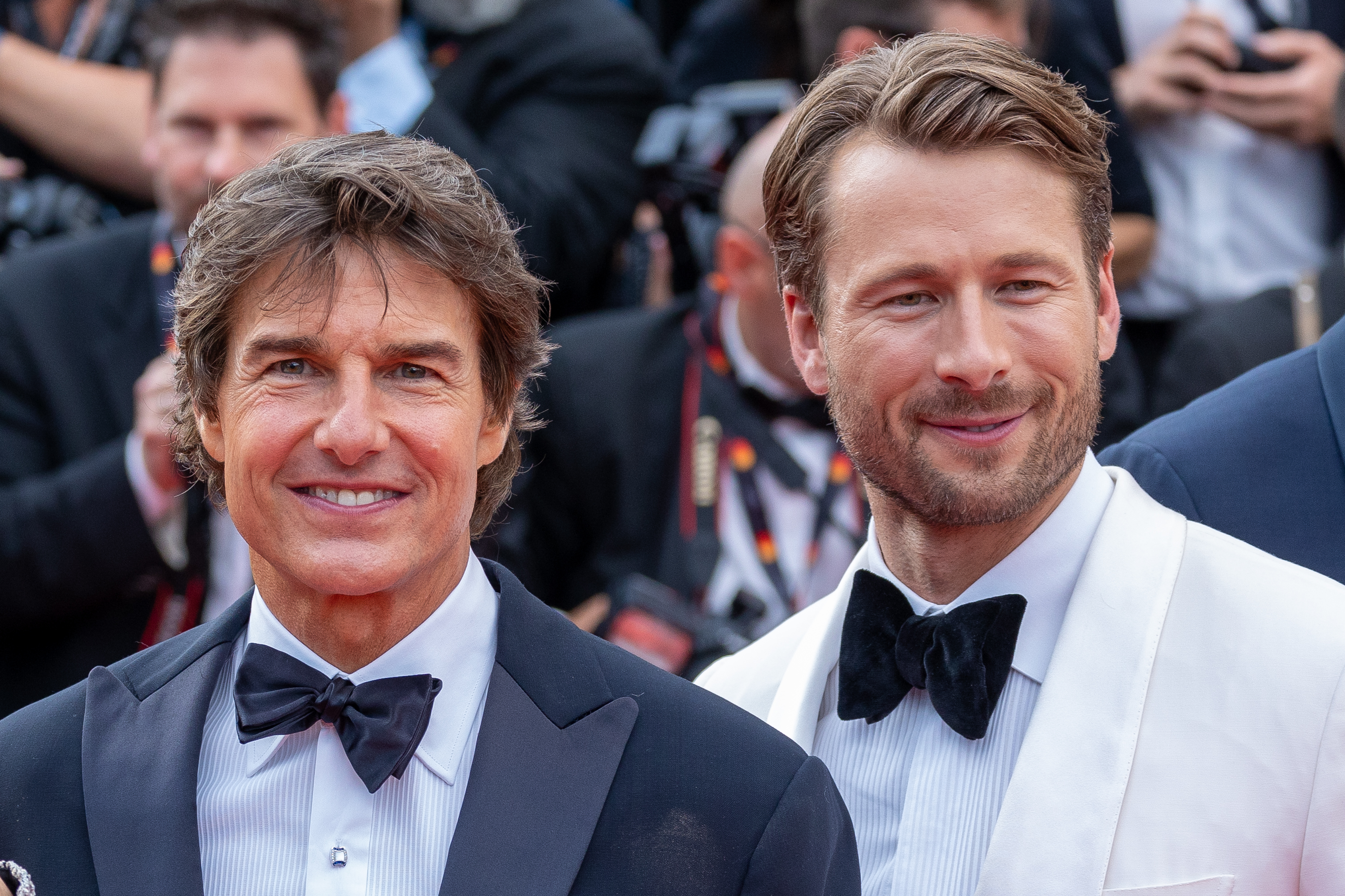 Tom Cruise and Glen Powell smiling on the red carpet, both wearing tuxedos with bow ties