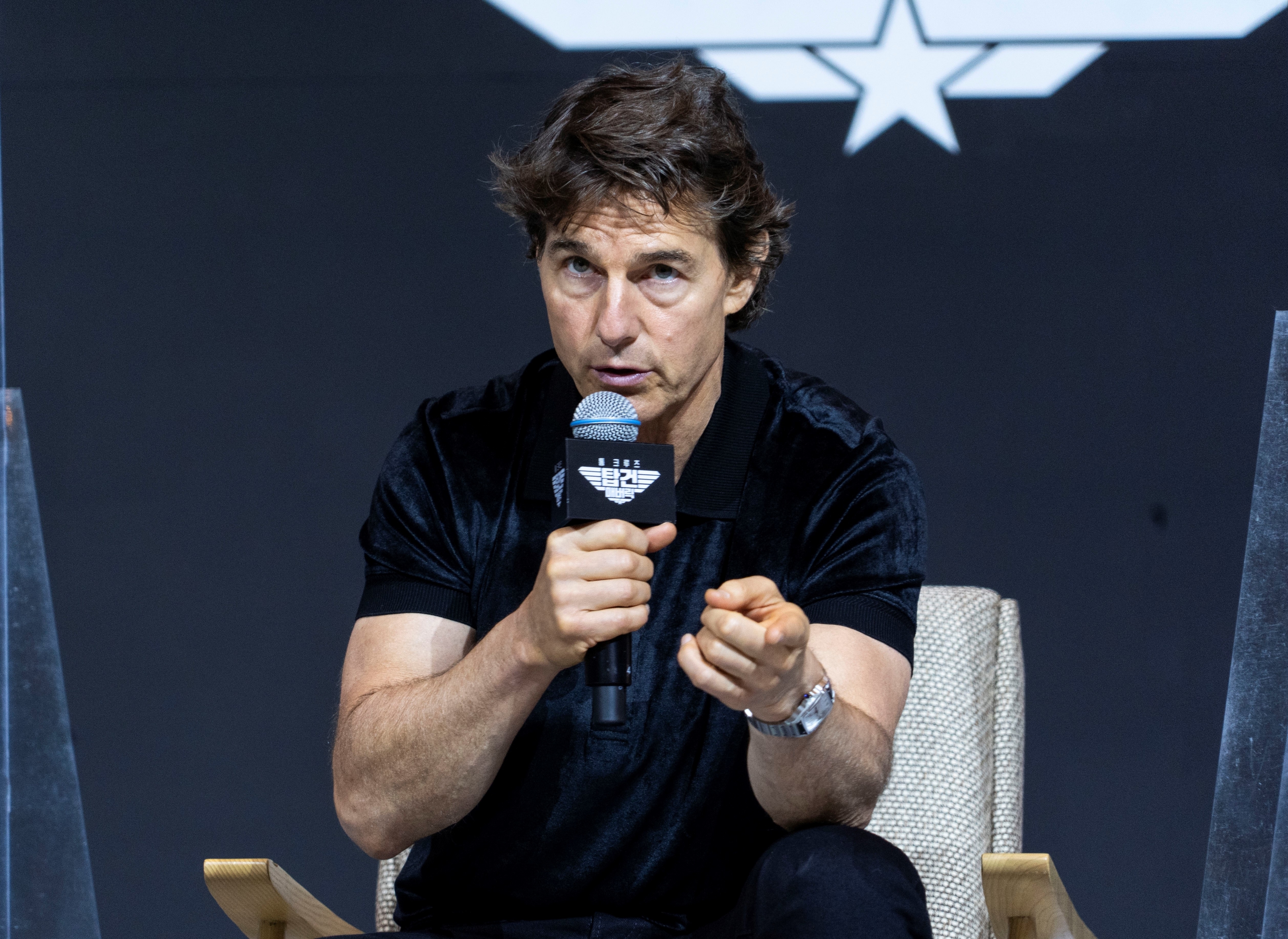 Tom Cruise speaks into a microphone while sitting in a chair, gesturing with his hand, during a public event