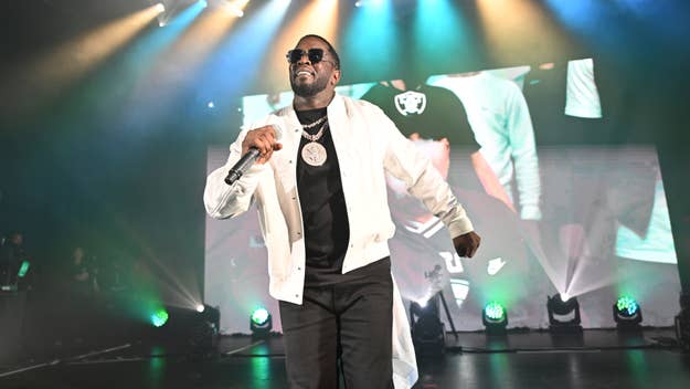 A man is performing on stage, holding a microphone. He is wearing sunglasses and a white jacket. Bright stage lights are in the background