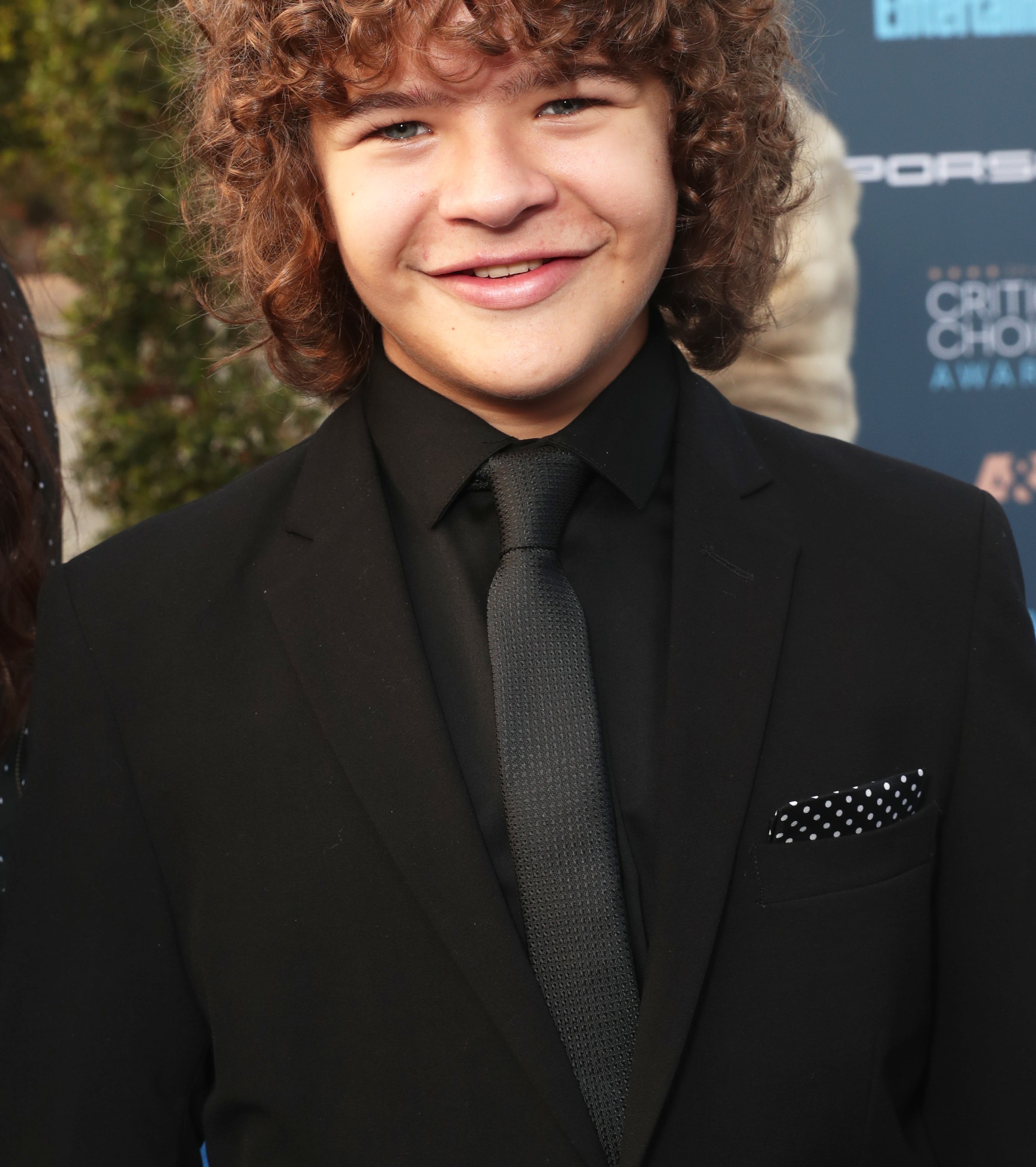 Gaten Matarazzo smiles while wearing a black suit and tie at an event