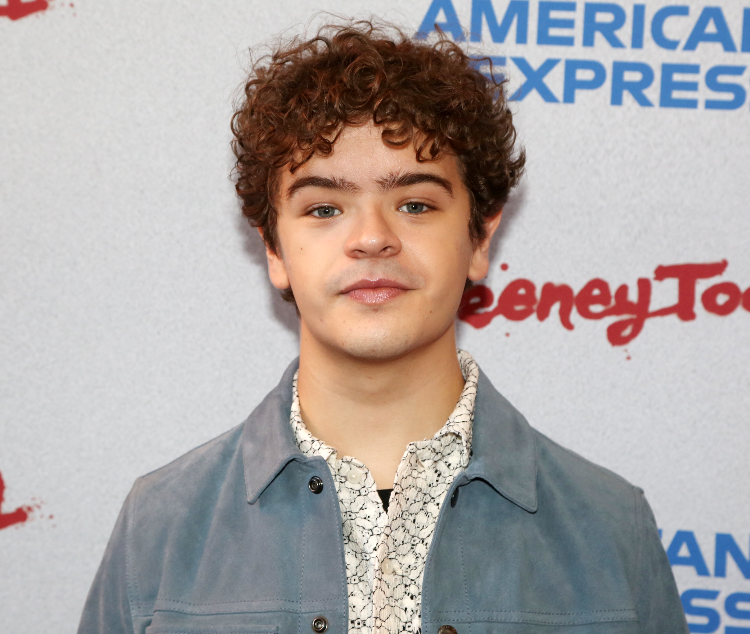 Gaten Matarazzo at an event, wearing a patterned shirt and a blue jacket, standing in front of a backdrop with the words &quot;American Express&quot; and &quot;Disney Toon.&quot;