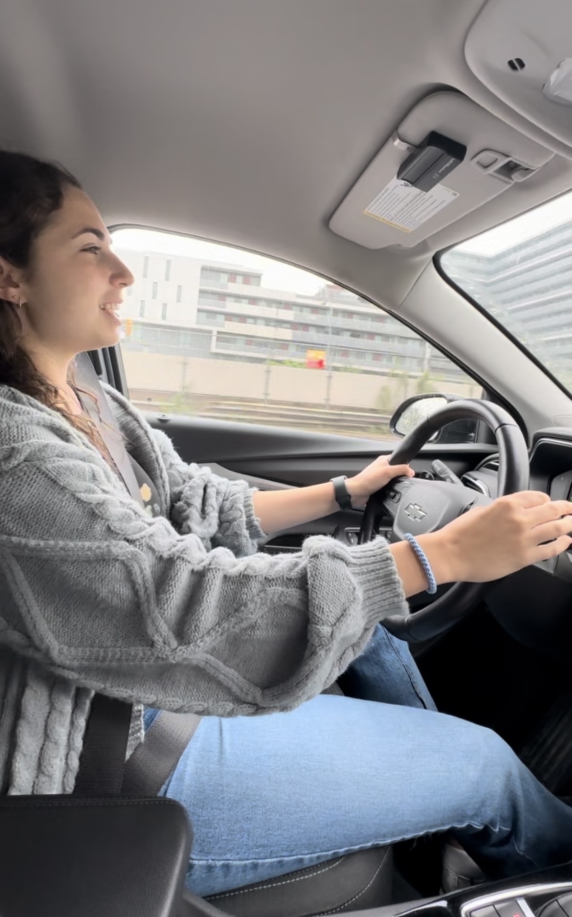 A person wearing a grey cardigan and jeans is smiling while driving a car. The urban landscape is visible through the windows