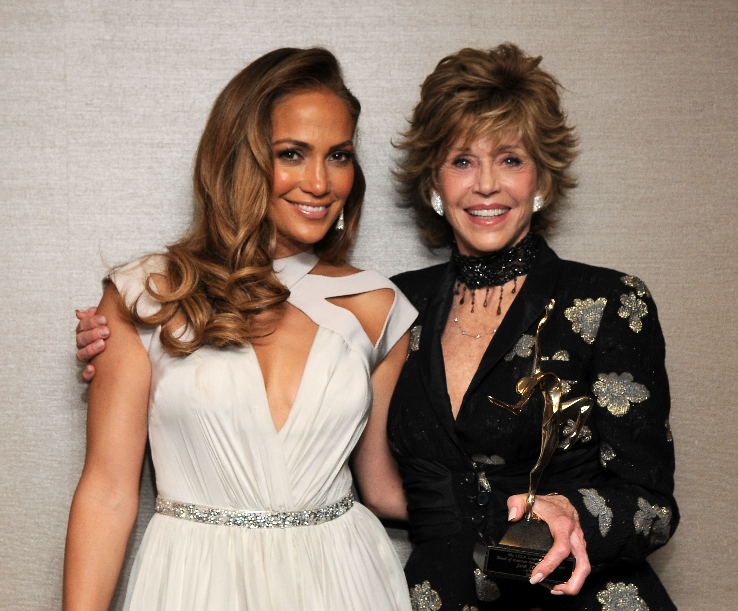 Jennifer Lopez in a gown and Jane Fonda in a floral dress, holding an award, pose together, smiling at an event