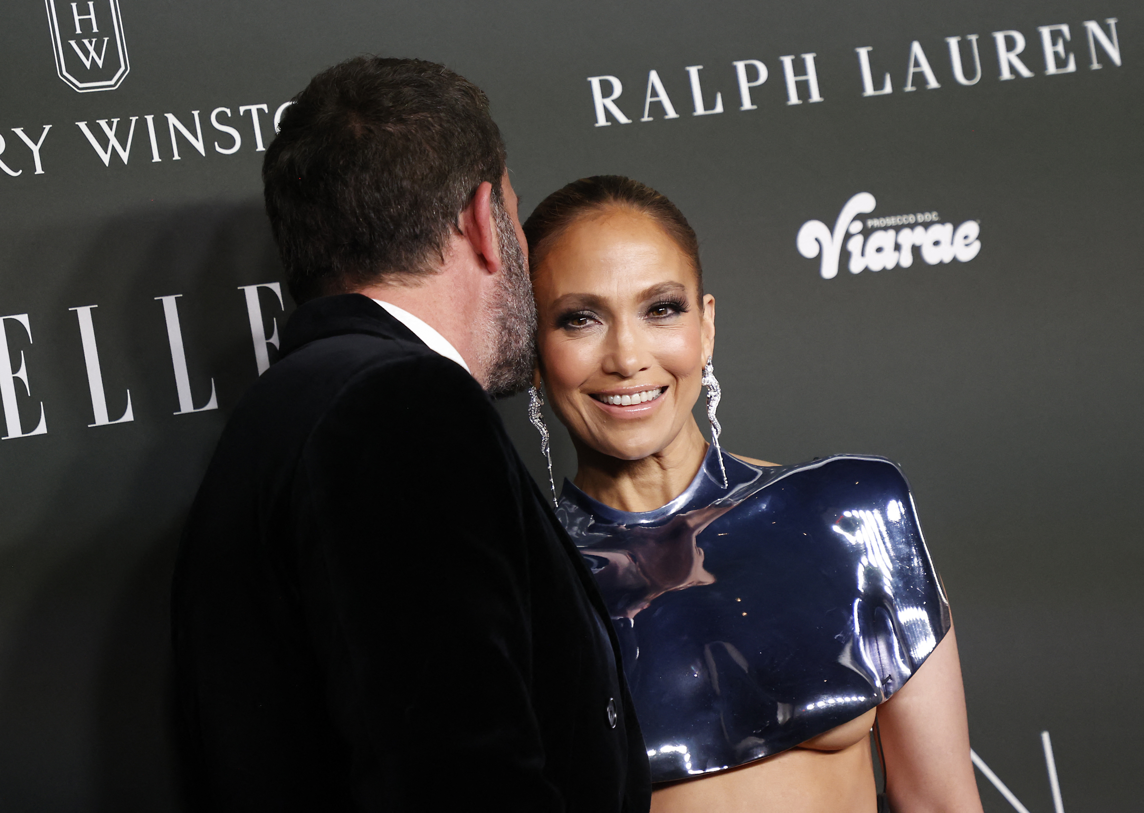 Jennifer Lopez in a sleek metallic outfit, smiles while a man whispers to her on a red carpet at an event