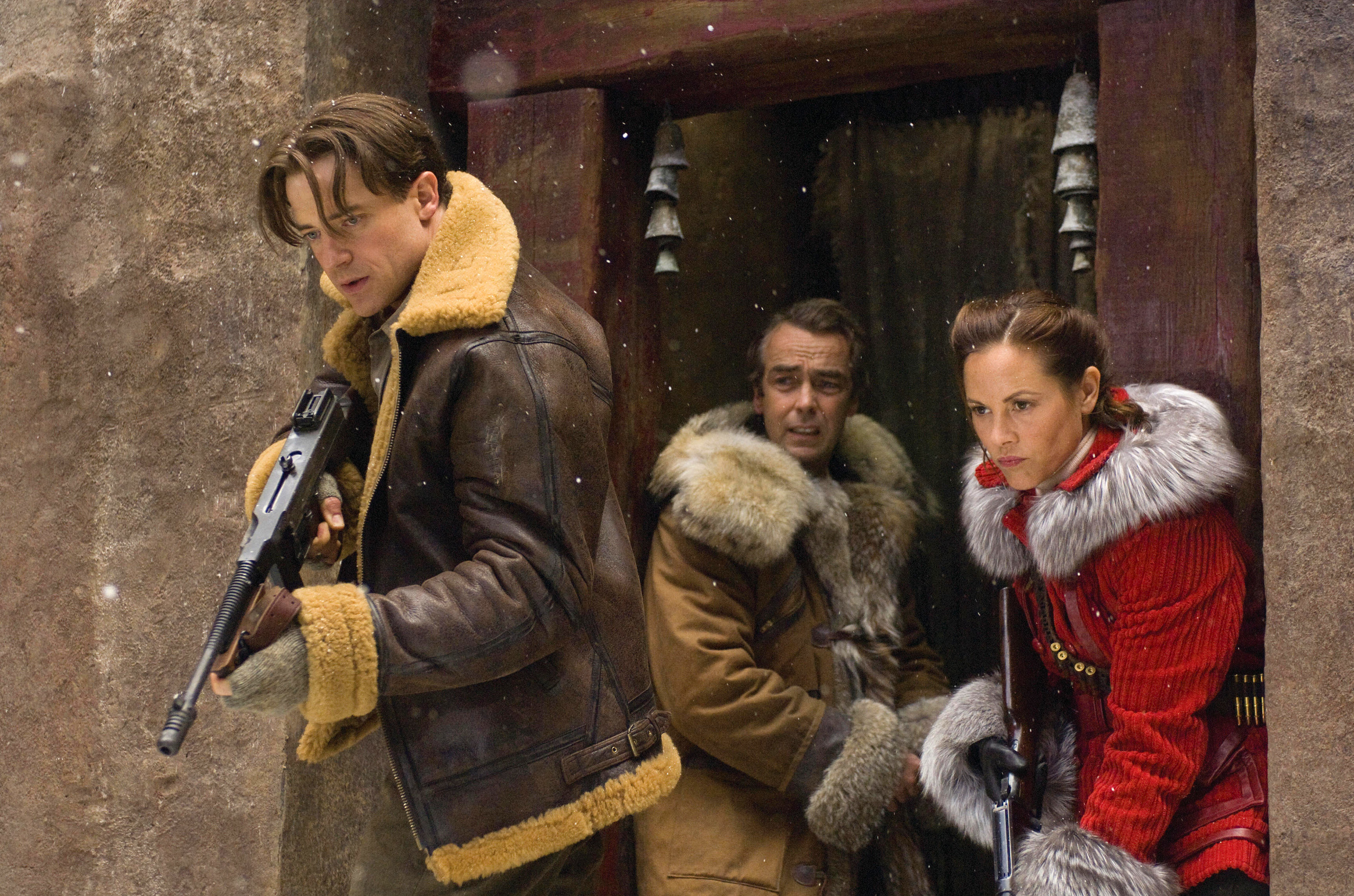 Brendan Fraser, John Hannah, and Maria Bello in warm, fur-lined jackets, holding weapons, standing in a snow-covered ancient doorway in a scene