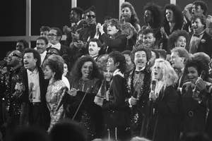 Lionel Richie, Diana Ross, Michael Jackson, Stevie Wonder, and other artists performing in a large group on stage, all singing and holding microphones