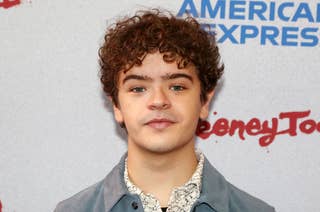Gaten Matarazzo at an event, wearing a patterned shirt and a blue jacket, standing in front of a backdrop with the words 
