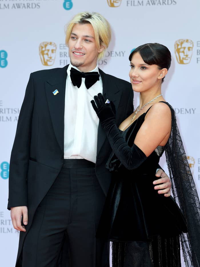 Jake Bongiovi in a tuxedo and Millie Bobby Brown in a dress with gloves, pose together on the red carpet at the BAFTA Film Awards