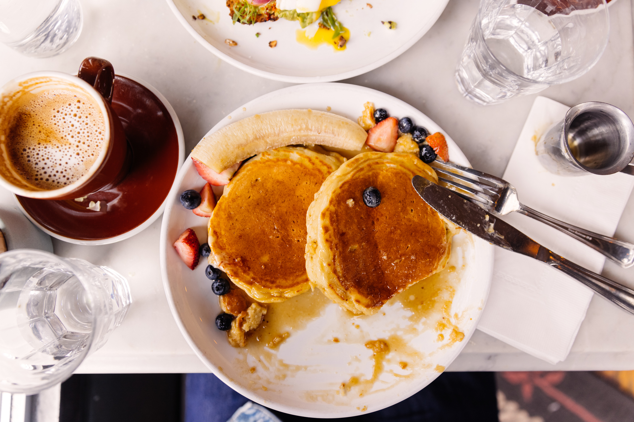 A plate with pancakes, sliced strawberries, bananas, and blueberries, accompanied by a cup of coffee and a glass of water on a table