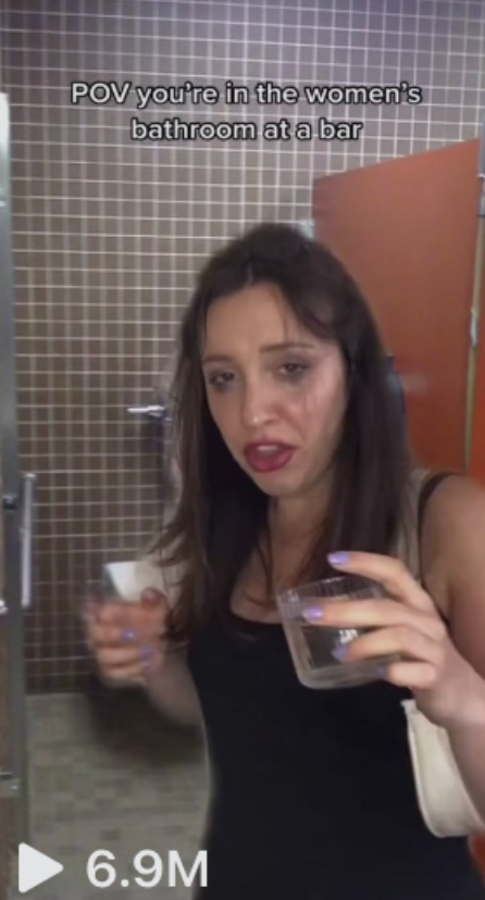 A woman is holding two drinks in a bar's women's restroom. Text reads, "POV you're in the women's bathroom at a bar." The video has 6.9M views