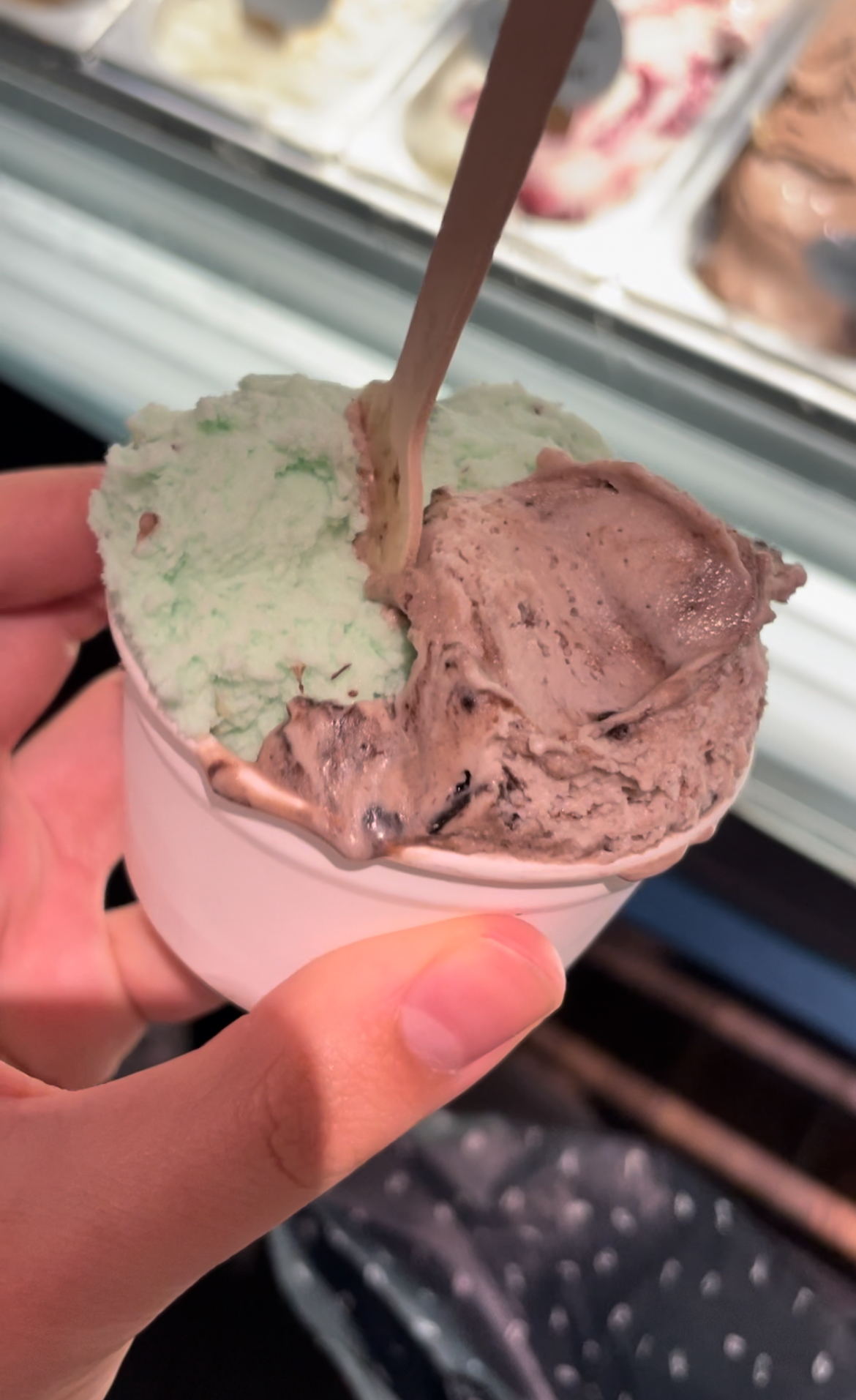A hand holding a cup of mint and chocolate ice cream with a plastic spoon, with more ice cream options visible in the background