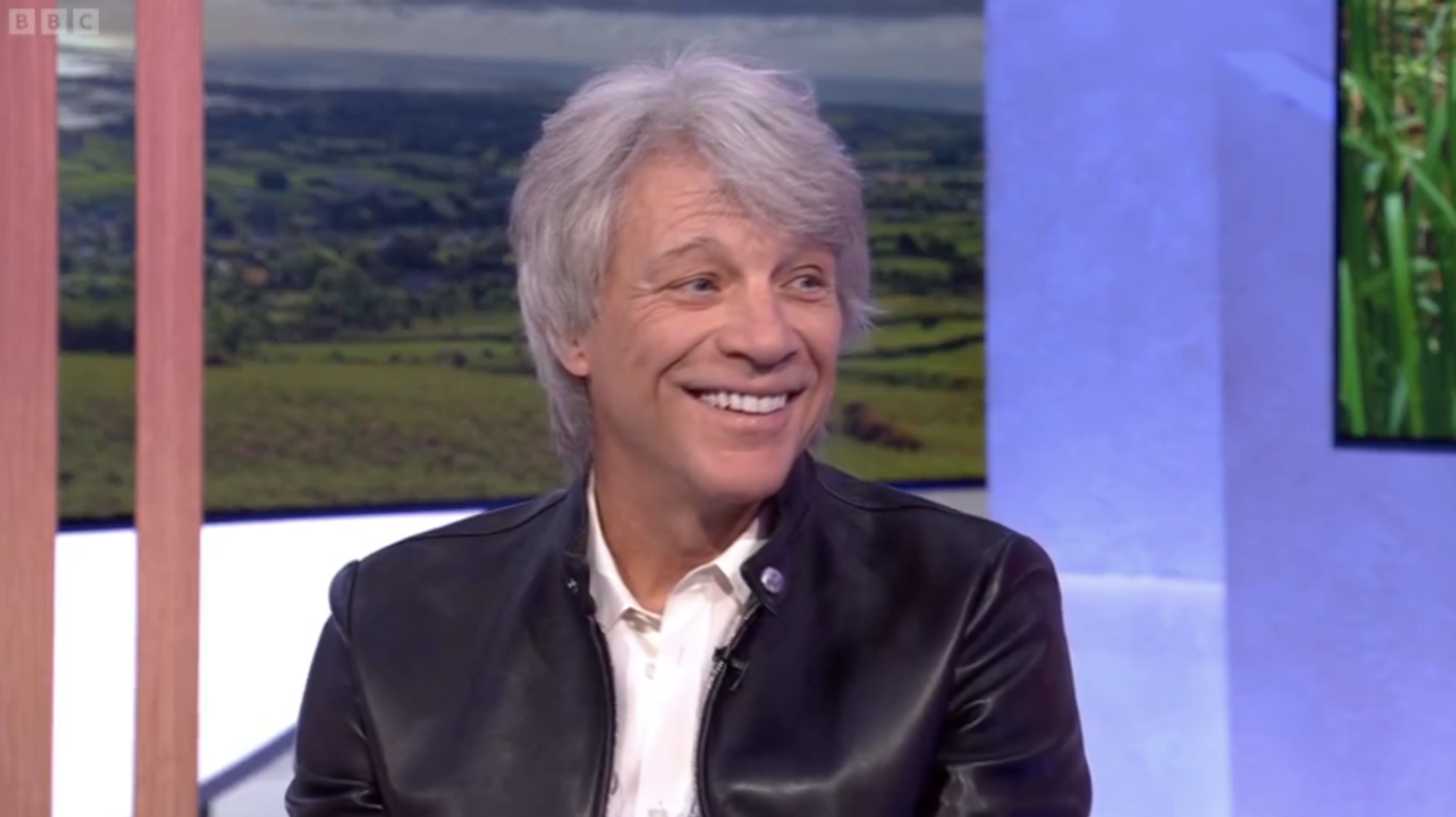 Jon Bon Jovi is smiling, wearing a black leather jacket and white shirt, during an interview on BBC