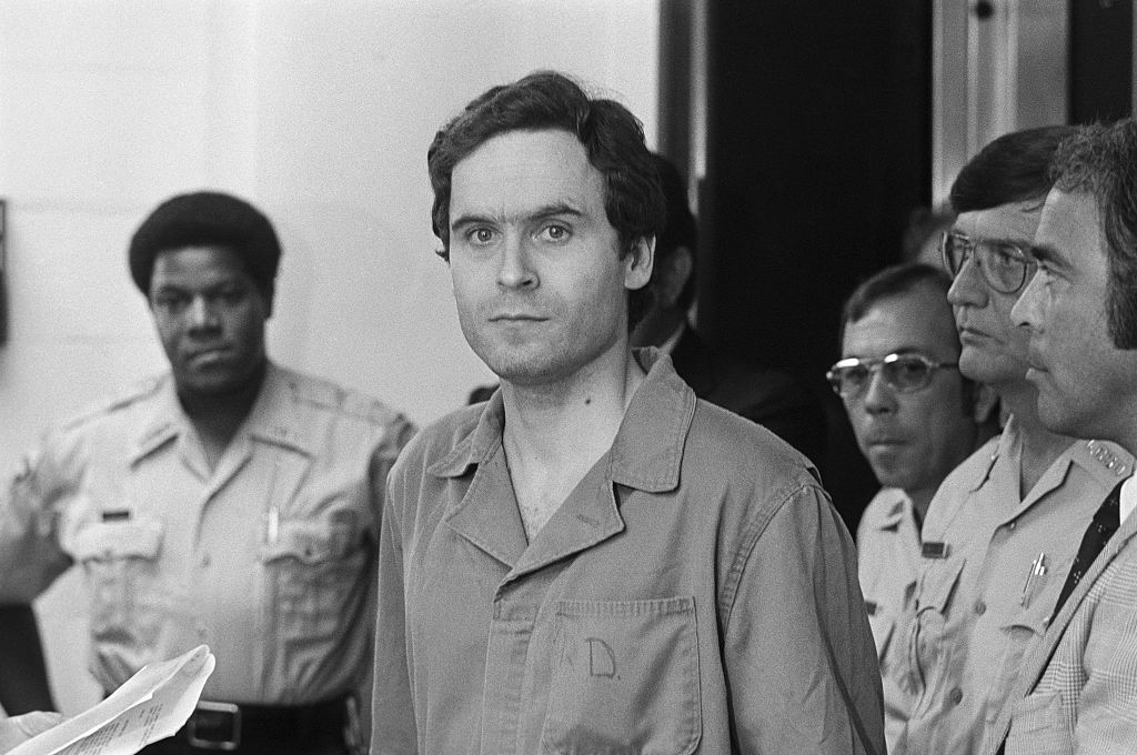 Ted Bundy is escorted by police officers while wearing a prison uniform in a black and white photograph