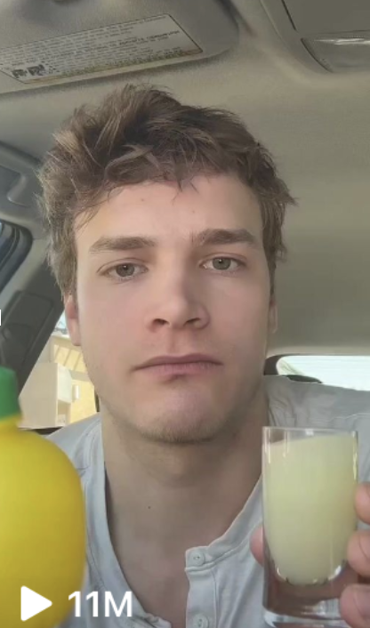 A man with short, tousled hair sits in a car holding a small glass of yellow liquid, with a lemon juice bottle beside him. The video has 11 million views