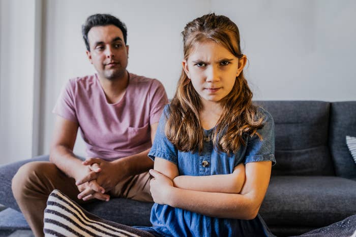 An upset young girl with folded arms sits on a couch, while a man in a pink shirt and brown pants looks at her with concern
