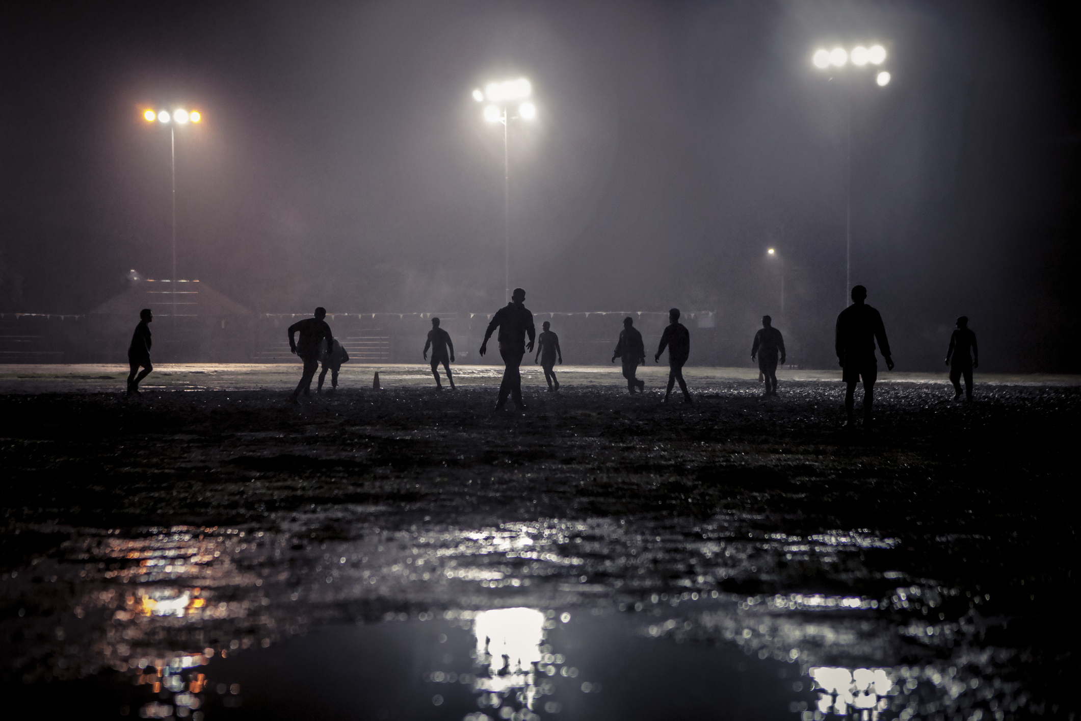 People playing soccer on a field at night under bright lights, with their silhouettes reflected in puddles on the ground